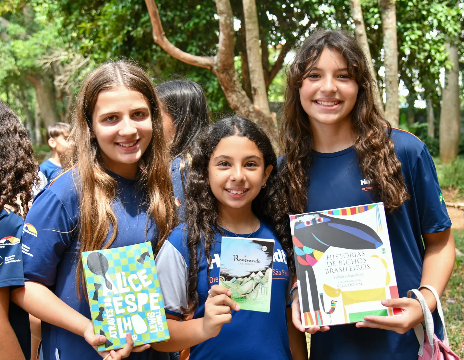 Alunas do fundamental 2 abraçadas em um espaço verde aberto e segurando livros nas mãos